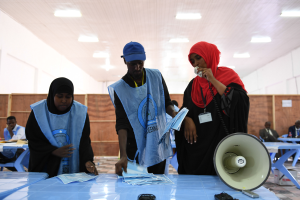Eine Gruppe von Menschen steht um einen Tisch mit einem Megafon und Papieren, während andere im Hintergrund eines Wahllokals mit beleuchteter Decke sitzen oder stehen.