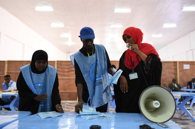 Eine Gruppe von Menschen steht um einen Tisch mit einem Megafon und Papieren, während andere im Hintergrund eines Wahllokals mit beleuchteter Decke sitzen oder stehen.