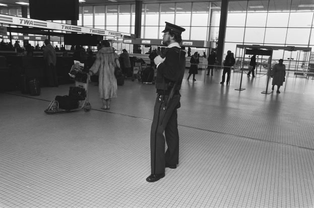 Polizeibeamter in Uniform mit Waffe in einem Flughafen-Terminal umgeben von Menschen, Gepäckwagen und Flughafen-Infrastruktur.