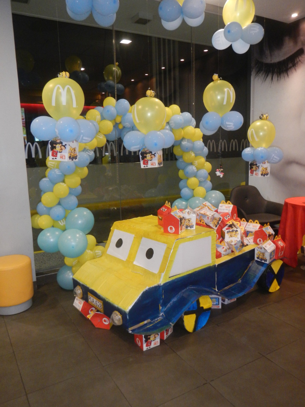 Eine McDonald's-Geburtstagsfeier mit einem gelben und blauen Lastwagen, der mit Ballons und anderen Dekorationen geschmückt ist, einem rot gedeckten Tisch mit Gegenständen, Hockern, einer Glaswand und Deckenlampen in einer festlichen Atmosphäre.