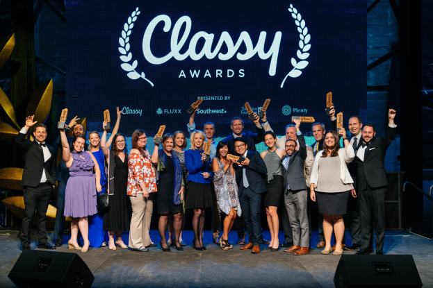 Eine Gruppe von Menschen auf einer Bühne, die lächeln und Preise halten, mit Lautsprechern unten und einem Bildschirm im Hintergrund, auf dem "Classy Awards 2019" angezeigt wird.