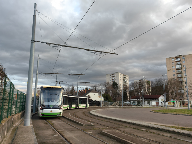 Grüne und weiße Straßenbahn auf Schienen neben hohen Gebäuden mit Strommasten und -leitungen, ein Zaun links und Bäume, Straßenlaternen und Gebäude im Hintergrund unter einem bewölkten Himmel.