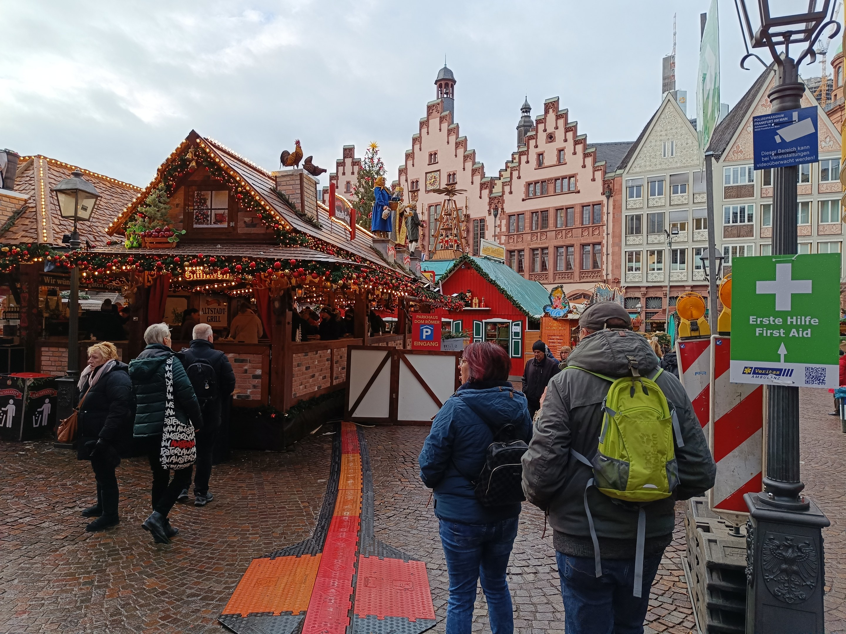Menschen, die auf einem Kopfsteinpflasterweg neben einem Weihnachtsmarkt in Nürnberg, Deutschland, gehen, mit Laternenmasten und Informationsschildern im Vordergrund und Gebäuden im Hintergrund.