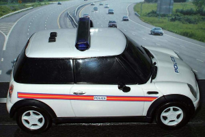 Ein weißes Polizeifahrzeug steht im Vordergrund, dahinter bewegen sich mehrere Fahrzeuge auf der Straße, umgeben von Gras und Bäumen auf beiden Seiten in einer ländlichen Umgebung.