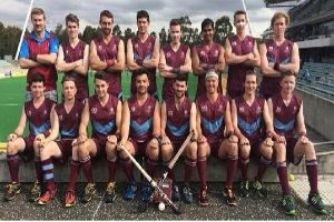 Eine Gruppe von Männern mit Hockey-Schlägern feiert auf einem Feld mit Bäumen und einem Stadion im Hintergrund.