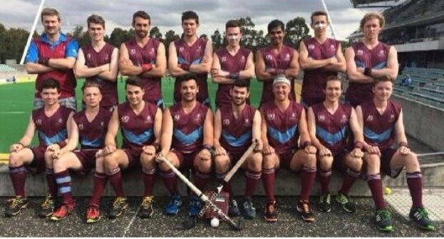 Eine Gruppe von Männern mit Hockey-Schlägern feiert auf einem Feld mit Bäumen und einem Stadion im Hintergrund.
