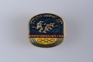 Ein runder Abzeichen mit dem Logo der Olympischen Spiele 1980, das zwei Läufer und den Text 'Olympic Games' darüber zeigt.
