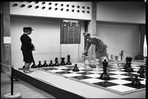 Zwei Frauen spielen Schach auf einem großen schwarz-weißen Brett vor einer Wand mit einem Brett daran, mit auf dem Brett angeordneten Schachfiguren.