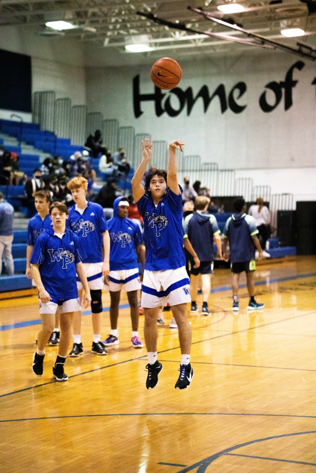 Eine Gruppe junger Männer spielt Basketball auf einem Indoor-Court mit einer Wand, die Text, Treppen, Geländer und Deckenleuchten zeigt, während Menschen in Sneakern zu sehen sind.