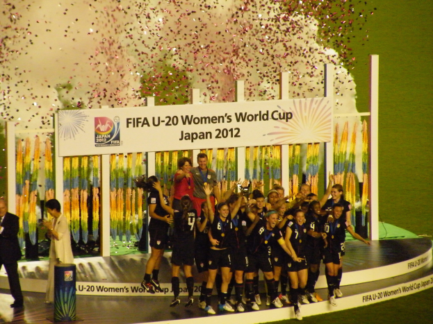 Gruppe von Frauenfußballspielerinnen in schwarzen Uniformen, die auf einer Bühne feiern, umgeben von einer Menge mit Kameras, mit einem Hintergrund aus Text, Bildern und Dekorationen auf Gras.