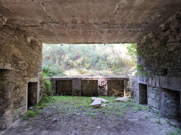 Alter Steinbau mit einem mittigen gewölbten Tunnel, umgeben von Mauern und Bäumen, und einem grasbewachsenen, steinigen Boden, der an einen Bunker erinnert.