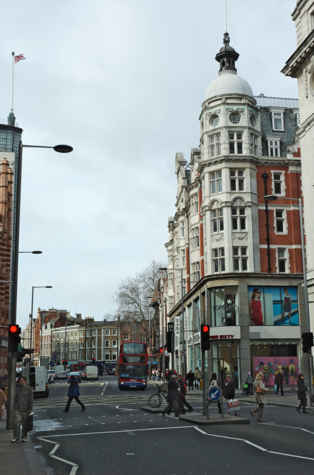 Ein roter Doppeldeckerbus fährt auf einer belebten Stadtstraße mit hohen Gebäuden, Laternen, Ampeln und Menschen auf dem Gehweg sowie Bäumen im Hintergrund und einem klaren blauen Himmel.