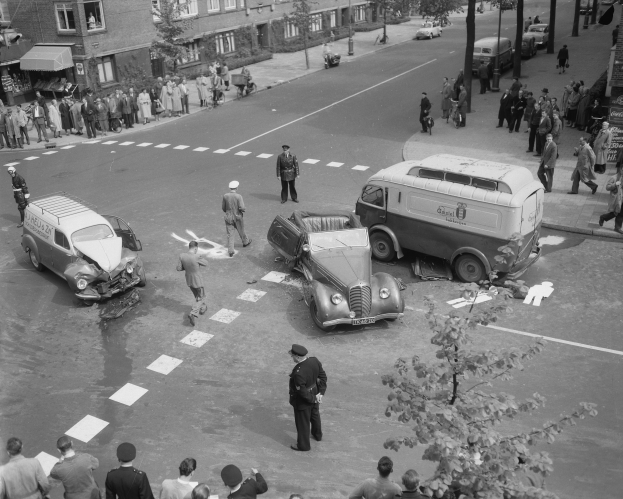 Schwarz-weiß-Szene eines Autounfalls an einer Straßenkreuzung mit einem Van, anderen Fahrzeugen und einer Gruppe von Schaulustigen; Gebäude, Bäume, Pfosten und Schilder im Hintergrund.