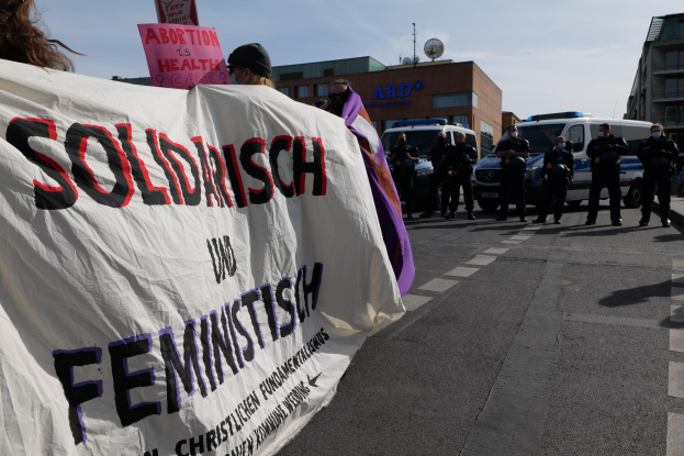 Eine Gruppe von Menschen marschiert mit einem Banner, auf dem 'Solidarität und Feminismus' steht, eine Straße entlang, mit parkenden Fahrzeugen am Straßenrand und Gebäuden im Hintergrund unter einem klaren blauen Himmel.