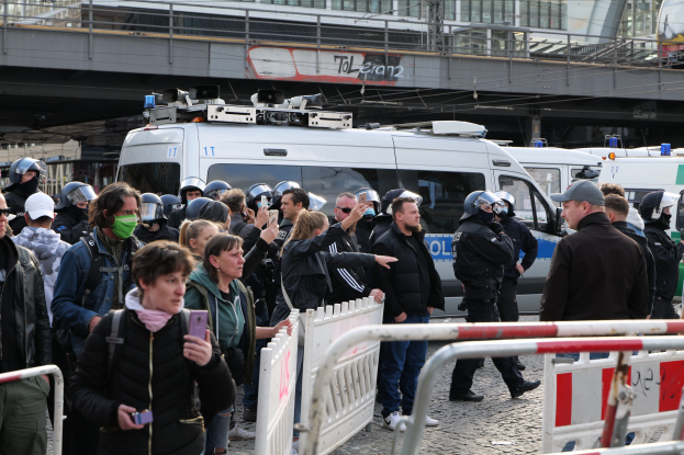 Eine Gruppe von Menschen steht vor Polizeiwagen in Berlin, einige tragen Helme und halten Telefone, mit Barrieren im Vordergrund und einer Brücke und Gebäuden im Hintergrund.