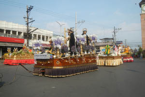 Ein Paradewagen mit Menschen darauf, umgeben von Gebäuden, Strommasten, Laternenmasten, Bäumen und einem Uhrenturm im Hintergrund unter einem bewölkten Himmel.