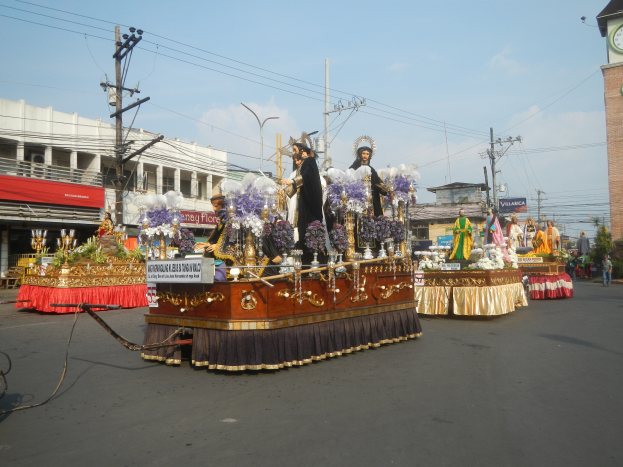 Ein Paradewagen mit Menschen darauf, umgeben von Gebäuden, Strommasten, Laternenmasten, Bäumen und einem Uhrenturm im Hintergrund unter einem bewölkten Himmel.