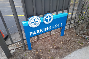 Ein blau-weißes Schild mit der Aufschrift "Parkplatz 1" steht vor einem Metallzaun, im Hintergrund ist eine Straße zu sehen und rechts ein Baum.