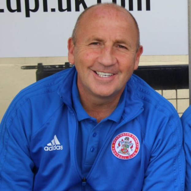 Zwei Männer in einem Raum, einer in einer blauen Jacke, der neben einem anderen Mann lächelt, neben einer Wandtafel im Hintergrund.