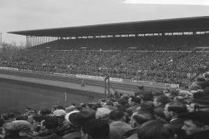 Schwarzes und weißes Foto eines vollen Stadions mit Zuschauern, die ein Fußballspiel beobachten, einige stehen und einige sitzen, mit Bannern, Pfählen, einer Hütte, Bäumen, einem Turm und einem bewölkten Himmel im Hintergrund.