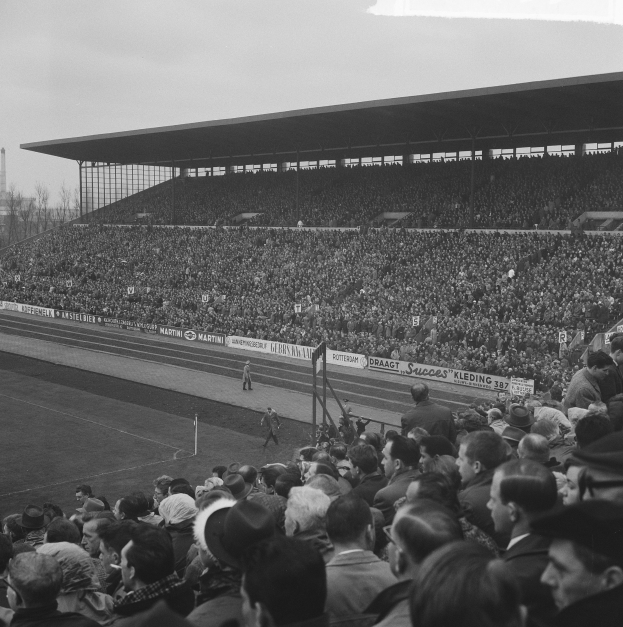 Schwarzes und weißes Foto eines vollen Stadions mit Zuschauern, die ein Fußballspiel beobachten, einige stehen und einige sitzen, mit Bannern, Pfählen, einer Hütte, Bäumen, einem Turm und einem bewölkten Himmel im Hintergrund.