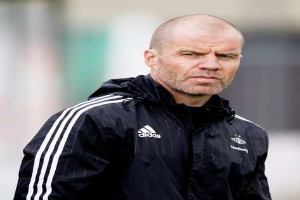 Glatzköpfiger Mann in einer blauen Jacke mit weißen Streifen und weißem Kragen, der auf einem Fußballfeld steht und die Hände verschränkt hat.