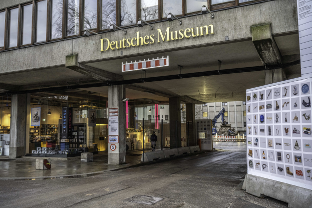 Außenansicht des Deutschen Museums in Berlin, mit einem großen Gebäude mit Glasfenstern, Säulen und einer Namensplakette, neben einer informativen Displaytafel, mit Bäumen, anderen Gebäuden und dem Himmel im Hintergrund.