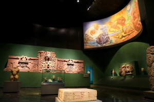 Eine Museumsausstellung im Anthropologischen Museum von Mexiko-Stadt, die verschiedene Artefakte zeigt, darunter Skulpturen, Gegenstände auf Tischen, eine Texttafel, eine Wandmalerei und Deckenbeleuchtung.
