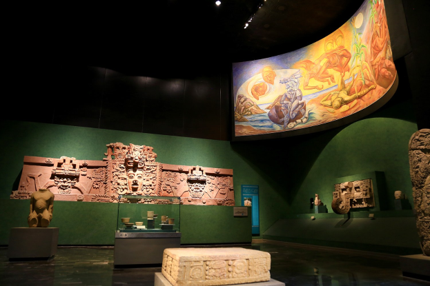 Eine Museumsausstellung im Anthropologischen Museum von Mexiko-Stadt, die verschiedene Artefakte zeigt, darunter Skulpturen, Gegenstände auf Tischen, eine Texttafel, eine Wandmalerei und Deckenbeleuchtung.