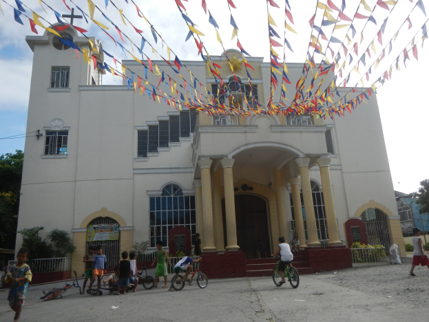 Eine Gruppe von Menschen, die auf Fahrrädern vor einer weißen Kirche mit architektonischen Details wie Fenstern, Säulen, Bögen, Türen und einem Kreuzsymbol steht, umgeben von Pflanzen, einem Zaun, einer Fahne, Bäumen und einem bewölkten Himmel, mit bunten Fahnen, die an einem Seil befestigt sind.