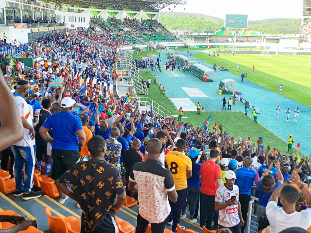 Eine große Menschenmenge versammelt sich vor einem eingezäunten Fußballfeld mit Hügeln und einem blauen Himmel im Hintergrund und wartet gespannt auf das Spiel der 2019 African Cup of Nations.
