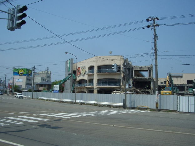 Stadtstraße mit Fahrzeugen, Strommasten, Laternen und einer Ampel auf der linken Seite, Gebäuden und Bäumen im Hintergrund und einem teilweise abgerissenen Gebäude im Vordergrund.