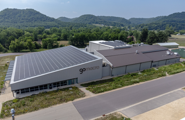 Luftaufnahme des Go Macro Solar Power Plant mit Solarmodulen auf dem Dach, umgeben von Grünflächen und einer Straße, mit Hügeln und einem klaren Himmel im Hintergrund.