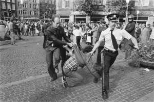 Ein Schwarz-Weiß-Bild eines Mannes, der von zwei Polizisten auf einer Straße mit vielen Menschen, Bäumen, Gebäuden, Laternen, Ampeln und Topfpflanzen im Hintergrund und einem Mannenschuh im Vordergrund gezogen wird.