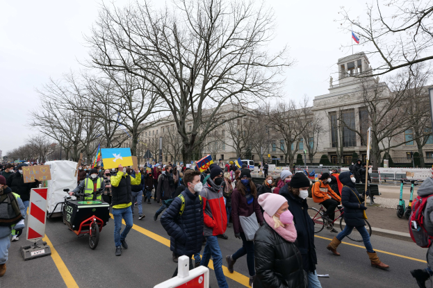 Eine große Gruppe von Menschen nimmt an einer Protestmarsch in Washington, D.C. am 21. Januar 2020 teil, geht eine Straße entlang, einige halten Plakate und Schilder, andere fahren Fahrráder, mit Bäumen, Schildern und einem klaren blauen Himmel im Hintergrund.