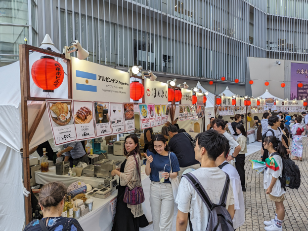 Eine Gruppe von Menschen auf einem Essensfest in Japan, die um einen Tisch mit Essen stehen, einige tragen Taschen und halten Gl├Ąser, mit Schildern, Lichtern und Laternen an der Decke und einem Geb├Ąude im Hintergrund.