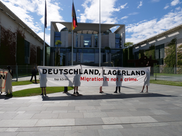 Eine Gruppe von Menschen mit einem Transparent, auf dem "Deutschland, Lagerland Migration ist kein Verbrechen" steht, vor einem Gebäude mit Fenstern, umgeben von Gras, einem Metallzaun, Pflanzen, Bäumen und Fahnen mit Stäben, unter einem bewölktem Himmel.