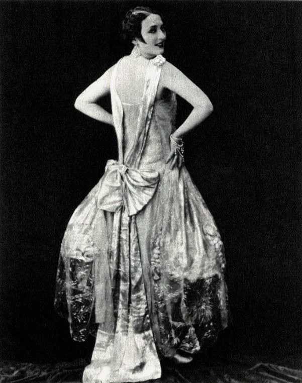 Eine Frau in einem Kleid im Stil der 1920er Jahre mit hohem Kragen, langen Ärmeln und einem weiten Rock steht selbstbewusst, ihr Haar ist elegant hochgesteckt und das Make-up natürlich.