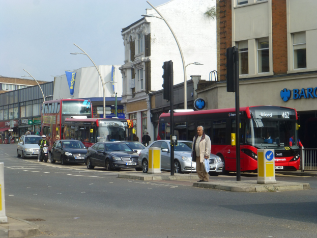 Ein roter Doppeldeckerbus fährt eine von hohen Gebäuden gesäumte Stadtstraße entlang, umgeben von Fußgängern, einem Radfahrer, Ampeln, Laternen und Texttafeln.