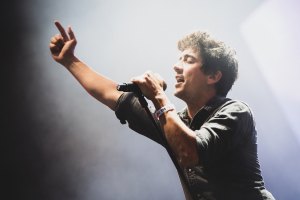 Ein Mann in schwarzem Hemd und Jeans singt in ein Mikrofon auf einer hell erleuchteten Bühne, die Augen geschlossen, während das Publikum applaudiert.