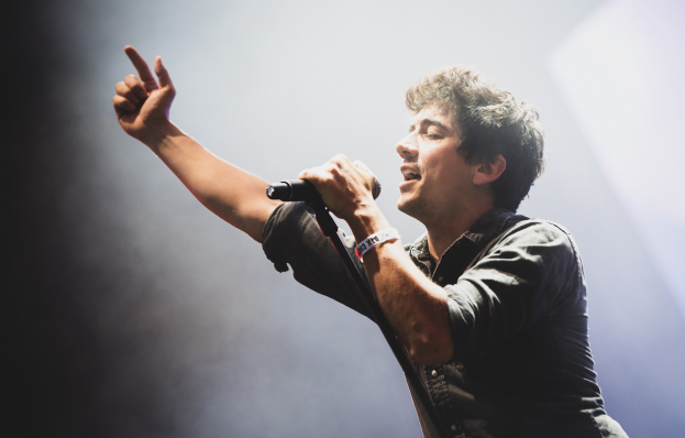 Ein Mann in schwarzem Hemd und Jeans singt in ein Mikrofon auf einer hell erleuchteten Bühne, die Augen geschlossen, während das Publikum applaudiert.