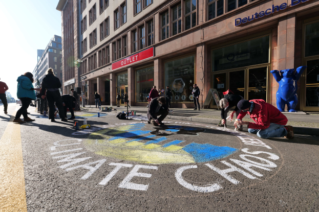 Menschen sitzen vor einem Gebäude während einer Klimaprotest in Berlin, umgeben von Flaschen und anderen Gegenständen mit Bäumen und einem klaren blauen Himmel im Hintergrund.