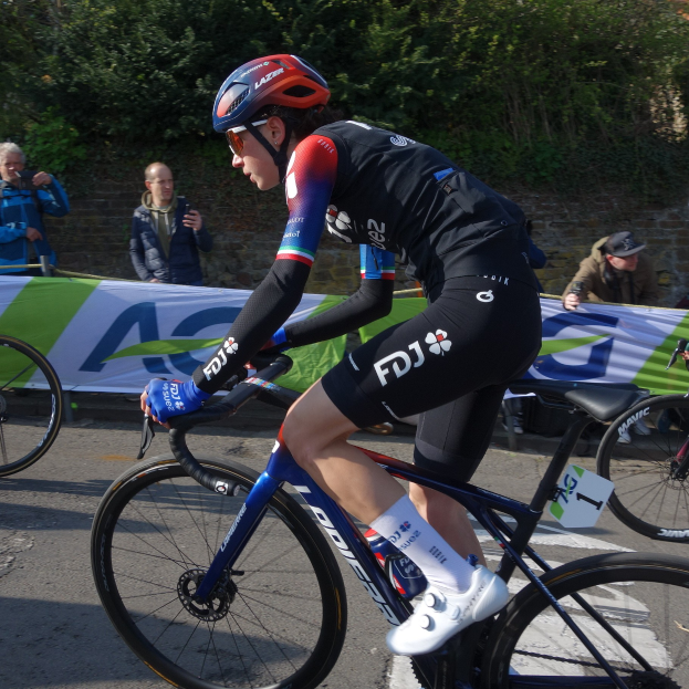 Professionelle Radsportlerin Katie Archibald mit Helm, Schutzbrille, Handschuhen und Schuhen auf einem Fahrrad die Straße entlangfahrend, vorbei an einer Menschenmenge, mit einem Banner, einer Wand, Bäumen und einem Gebäude im Hintergrund.