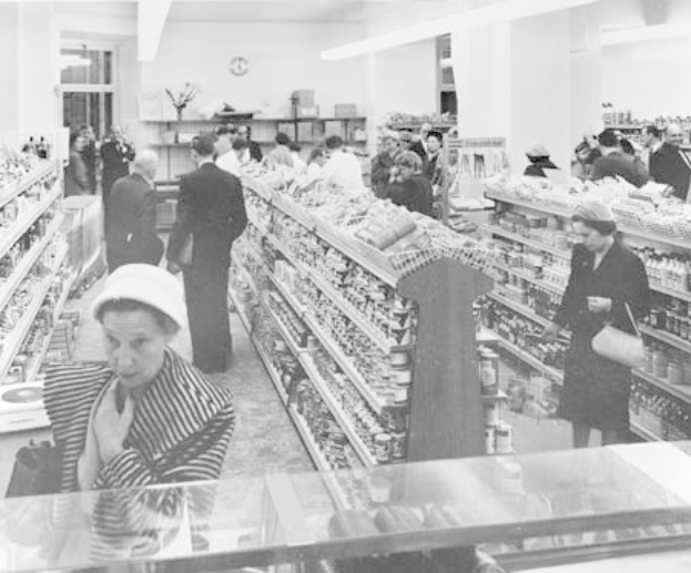 Schwarzes und weißes Foto von Menschen beim Einkaufen in einem Supermarkt mit Regalen voller Lebensmittel, einige halten Taschen, eine Uhr an der Wand und Deckenleuchten.