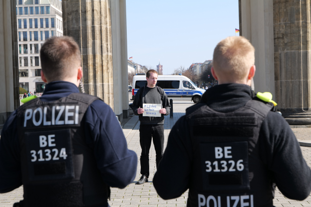 Drei Polizeibeamte stehen vor einem Gebäude mit einem Mann in der Mitte, der ein Papier hält, flankiert von Säulen, mit Fahrzeugen, Menschen, Bäumen und Gebäuden im Hintergrund.