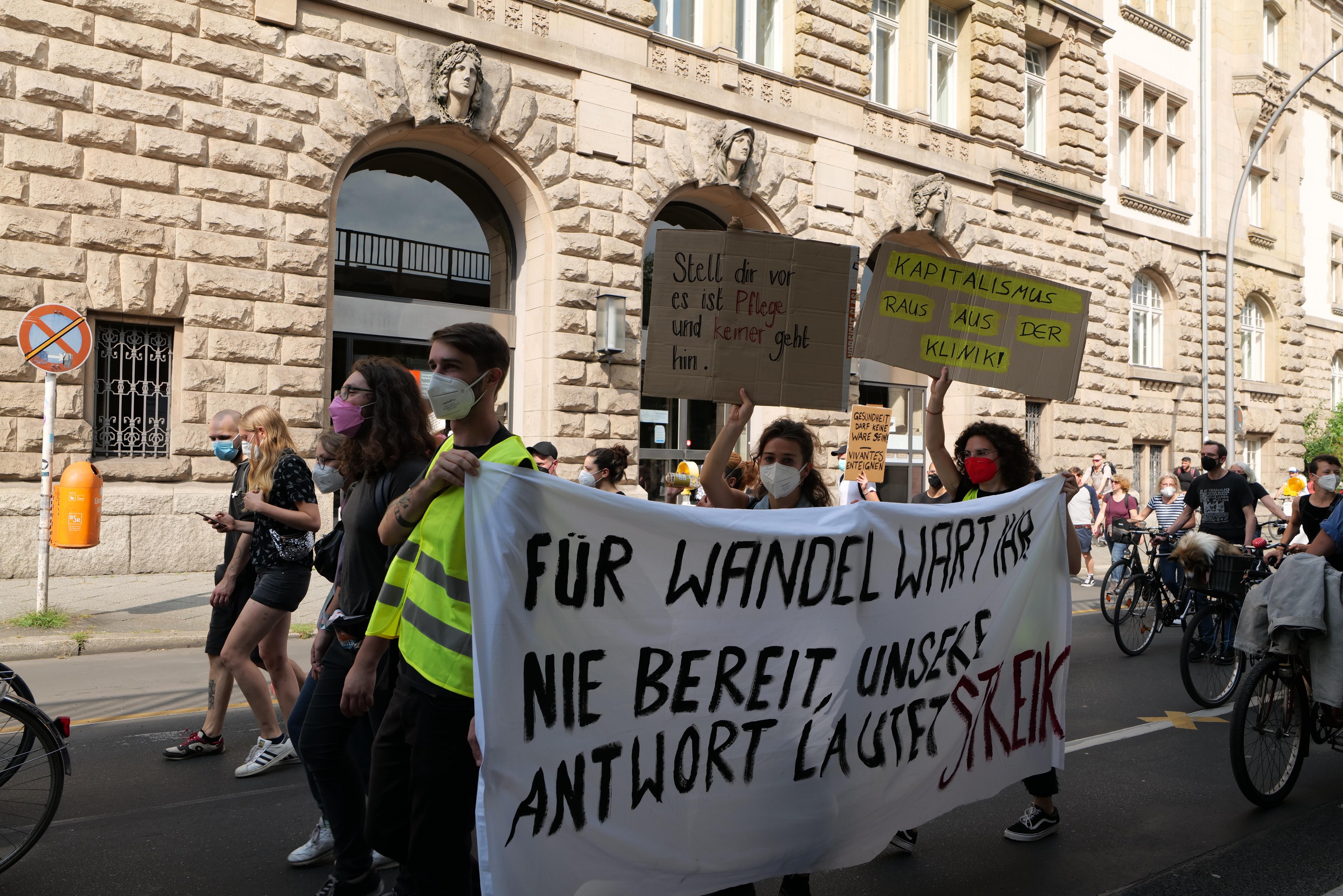 Eine Gruppe von Menschen marschiert bei einer Protestaktion gegen die Anti-Kriegs-Bewegung in Berlin, einige halten Schilder und andere fahren Fahrräder, im Hintergrund ist ein Gebäude mit Fenstern, Bögen, Säulen und Skulpturen, Bäumen und einem Laternenpfahl zu sehen.