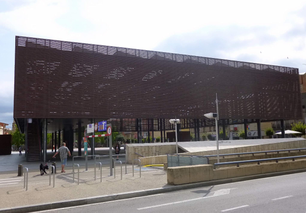 Großes Gebäude mit vielen Fenstern, neuer Stadtzentrumseingang, umgeben von Straßeninfrastruktur, Fahrzeugen, Fußgängern und Bäumen unter einem bewölkten Himmel.