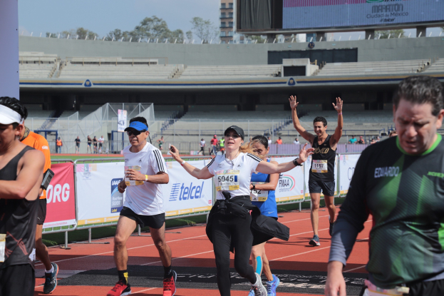 Gruppe von Läufern auf einer Stadionbahn mit Bannern, einem Zaun, einem Display, einem Gebäude, Bäumen und einem bewölkten Himmel; einige Läufer tragen Turnschuhe und Hüte.