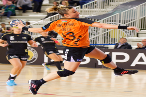 Eine Frau in einem orangen T-Shirt und schwarzen Shorts spielt Handball und hält einen Ball, mit Zuschauern und einem Banner mit der Aufschrift "Krp Slezak - Handball-Weltmeisterschaft 2019" im Hintergrund.