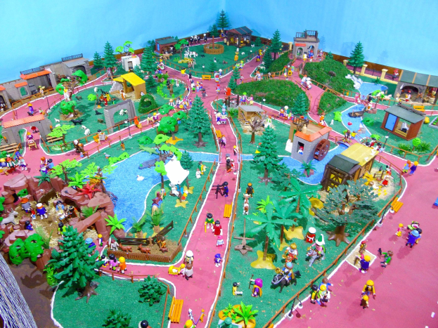 Ein Miniaturzoo-Modell mit Menschen bevölkert, umgeben von Bäumen, Häusern, Pflanzen, Gras, einem Zaun und anderen Gegenständen, mit einer Wand im Hintergrund und Text unten.